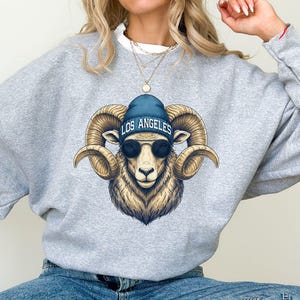 May include: Sport gray sweatshirt featuring a detailed illustration of a ram wearing a blue beanie with "LOS ANGELES" text and sunglasses. The ram has large curved horns and a detailed fur texture. The sweatshirt is a neutral color.