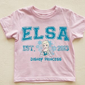 A pink t-shirt with the text "ELSA EST. 2013 DISNEY PRINCESS" printed on it, along with a depiction of the Disney princess Elsa.