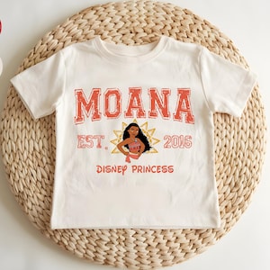 A white t-shirt with the text "Moana" and "Disney Princess" printed on it, along with a character illustration. The t-shirt is placed on a woven mat.