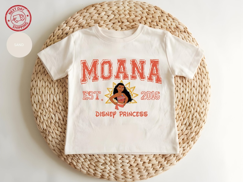 A white t-shirt with the text "Moana" and "Disney Princess" printed on it, along with a character illustration. The t-shirt is placed on a woven mat.