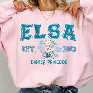A person wearing a pink sweatshirt with the text "ELSA EST. 2013 DISNEY PRINCESS" printed on it, along with a depiction of the Disney princess Elsa.