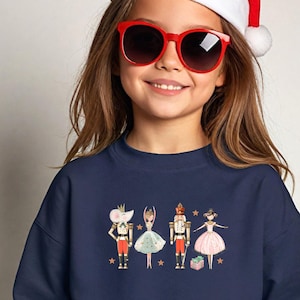 May include: Navy blue sweatshirt featuring a whimsical illustration of four Nutcracker characters. The design includes a mouse king, a ballerina in a light blue dress, a nutcracker soldier, and a ballerina in a pink dress. The word "Navy" is printed on the top right.