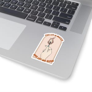 May include: A sticker featuring a hand holding a lit match. The match has a flame with red, orange, and yellow hues. The sticker has the text "YOU LIT A MATCH AND LET ME BURN" in a stylized font, with a light pink background.