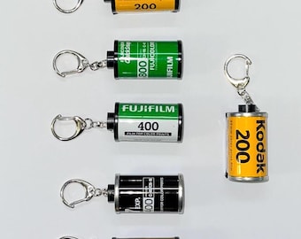 May include: A collection of film canister keychains. The keychains feature various film brands, including Kodak Gold, Fujifilm, and Kodacolor. Each keychain has a silver keyring attached to a miniature film canister with text and numbers.