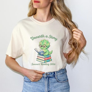 May include: Cream-colored t-shirt featuring a cartoon dinosaur wearing glasses and reading a book atop a stack of books. The text "Unearth a Story" and "Summer Reading 2026" are printed in green.