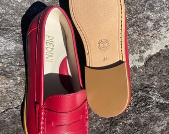 May include: Red leather loafers with a classic penny loafer design. The sole is visible, and the size is 39. The brand name "PIEDINI" is visible on the insole. The shoes are displayed on a textured, gray surface.