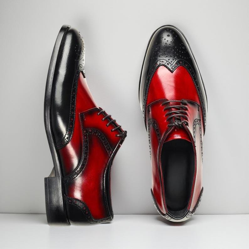 May include: A pair of two-tone leather dress shoes. The shoes feature a black toe cap and heel, with a vibrant red body. The shoes have a classic wingtip design with decorative perforations and are laced up with dark brown laces.
