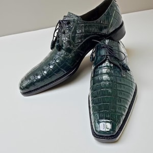 May include: A pair of dark green leather dress shoes with a textured, crocodile-like pattern. The shoes have a lace-up design with tassels and a square toe. The soles are black.