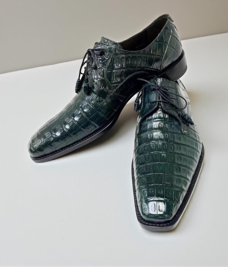 May include: A pair of dark green leather dress shoes with a textured, crocodile-like pattern. The shoes have a lace-up design with tassels and a square toe. The soles are black.