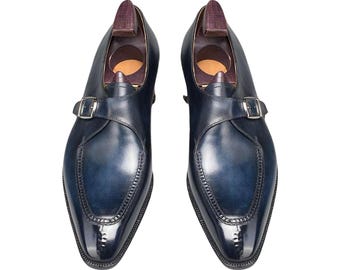 May include: A pair of navy blue leather dress shoes with a glossy finish. Each shoe features a silver buckle closure and a wooden shoe tree inside. The shoes have a pointed toe and decorative stitching.