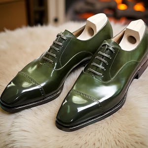 Op de afbeelding: Een paar gepolijste, donkergroene leren herenschoenen met een cap-toe ontwerp. De schoenen hebben grijze veters en worden getoond met houten schoenspanners erin. De schoenen staan op een lichtgekleurd, getextureerd oppervlak.