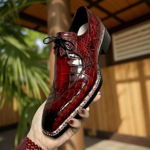 May include: A red leather dress shoe with a glossy finish and a textured, crocodile-like pattern. The shoe features a square toe, black laces, and a small heel. The shoe has a silver trim around the sole.