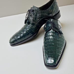 May include: A pair of dark green leather dress shoes with a textured, crocodile-like pattern. The shoes have a lace-up design with tassels and a square toe. The soles are black.