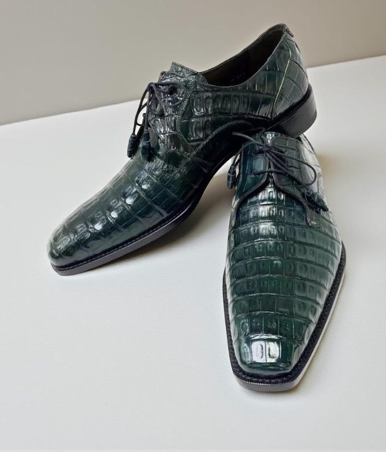 May include: A pair of dark green leather dress shoes with a textured, crocodile-like pattern. The shoes have a lace-up design with tassels and a square toe. The soles are black.
