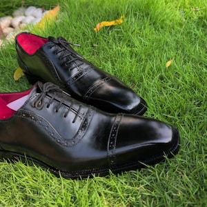 Puede incluir: Un par de zapatos oxford de cuero negro con dise&ntilde;o de puntera y detalles decorativos de brogue a lo largo de las costuras y los paneles de los ojales. Los zapatos tienen cordones negros y un forro interior rosa intenso visible.