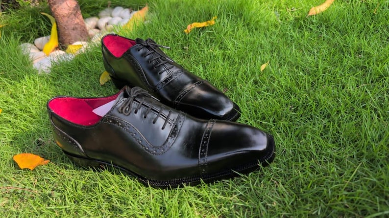 Puede incluir: Un par de zapatos oxford de cuero negro con dise&ntilde;o de puntera y detalles decorativos de brogue a lo largo de las costuras y los paneles de los ojales. Los zapatos tienen cordones negros y un forro interior rosa intenso visible.