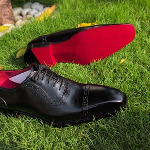 Puede incluir: Un par de zapatos oxford de cuero negro con puntera reforzada y detalles brogue decorativos, cierre de cordones y una suela de color rojo vibrante. El forro interior del zapato tambi&eacute;n es de color rojo brillante, contrastando con el exterior.