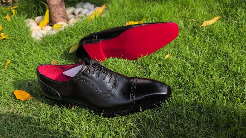 Puede incluir: Un par de zapatos oxford de cuero negro con puntera reforzada y detalles brogue decorativos, cierre de cordones y una suela de color rojo vibrante. El forro interior del zapato tambi&eacute;n es de color rojo brillante, contrastando con el exterior.
