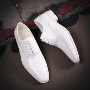 May include: A pair of white patent leather oxford shoes with a cap toe design, featuring a lace-up closure with matching white laces, a low block heel, and a smooth, glossy finish that reflects light across the surface of the footwear.