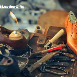 Puede incluir: Un zapato oxford de cuero color canela con detalles brogue y una horma de color claro en su interior, colocado sobre un banco de trabajo con diversas herramientas de zapatero, incluyendo martillos e instrumentos de metal. El texto ASLeatherGifts aparece arriba a la izquierda.