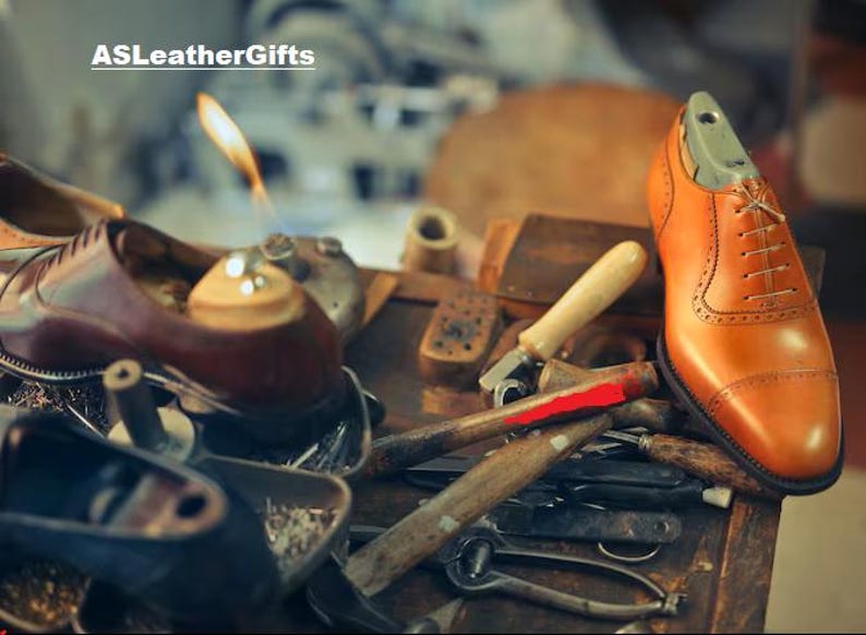 Puede incluir: Un zapato oxford de cuero color canela con detalles brogue y una horma de color claro en su interior, colocado sobre un banco de trabajo con diversas herramientas de zapatero, incluyendo martillos e instrumentos de metal. El texto ASLeatherGifts aparece arriba a la izquierda.