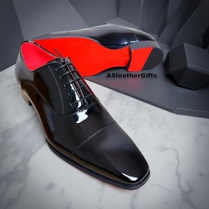 May include: Black patent leather dress shoes with red soles and interior. The shoes have a classic Oxford design with a lace-up closure. The image shows two shoes, one in focus and the other in the background. The brand name "ASleatherGifts" is visible.