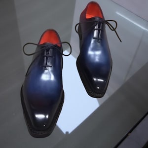 May include: A pair of two-tone dress shoes. The shoes transition from a deep navy blue to black at the toe, with a red interior. The shoes have a glossy finish, laces, and a classic Oxford design.