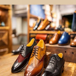 May include: Leather dress shoes in various colors, including black, brown, and burgundy, are displayed on a wooden surface. The text "ASLeatherGifts" is at the top of the image. Other shoes are visible on shelves in the background.