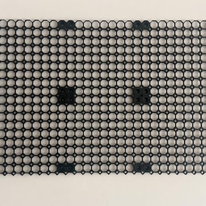 May include: A black plastic grid with a repeating pattern of circular openings. The grid is rectangular and has several small, solid black squares evenly spaced across its surface. The background is a plain, off-white color.