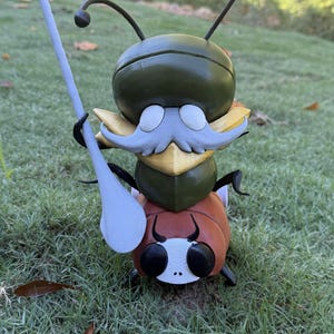 May include: A whimsical, 3D-printed insect figurine with a green head, yellow accents, and a brown body. It has large black eyes, a white mustache, and holds a light gray spear. The figure is set against a backdrop of green grass.