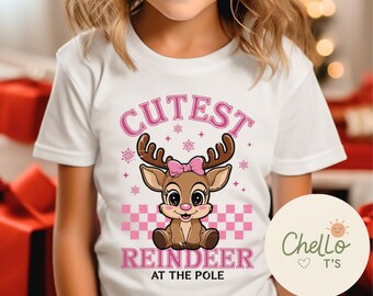 May include: White t-shirt featuring a cartoon reindeer with a pink bow, antlers, and the text "Cutest Reindeer at the Pole" in pink. The design includes pink snowflakes and a checkered pattern.