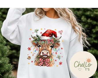 May include: White sweatshirt featuring a watercolor illustration of a Highland cow decorated for Christmas. The cow wears a Santa hat, a plaid scarf, and festive lights. The design includes snowflakes and the text "Chello T's".