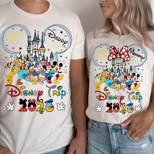Puede incluir: Camisetas beige a juego con un colorido dise&ntilde;o de Disney. El dise&ntilde;o incluye un castillo, fuegos artificiales y personajes de Disney. El texto "Disney Trip 2020" se muestra debajo. Una camiseta dice "Daddy" y la otra dice "Mommy".