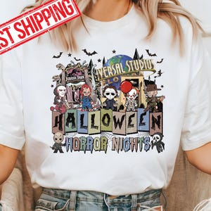 May include: A white t-shirt with a graphic print of a Halloween scene. The scene features a group of horror movie characters in front of a Universal Studios Halloween Horror Nights sign. The characters include Freddy Krueger, Jason Voorhees, Michael Myers, Pennywise, and Chucky. The text on the sign reads 'Universal Studios Halloween Horror Nights'. The t-shirt is being worn by a person with blue jeans.
