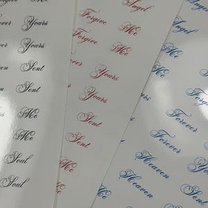 May include: Three sheets of white adhesive stickers featuring repeating words in elegant cursive script. The text includes Forgive Me, Yours, Sent, Me, Soul, Fallen Angel, Forever, Heaven, and Kiss, printed in black, red, and blue ink colors.