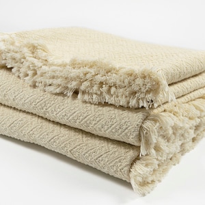 May include: A folded cream-colored throw blanket with a herringbone pattern and fringe trim.