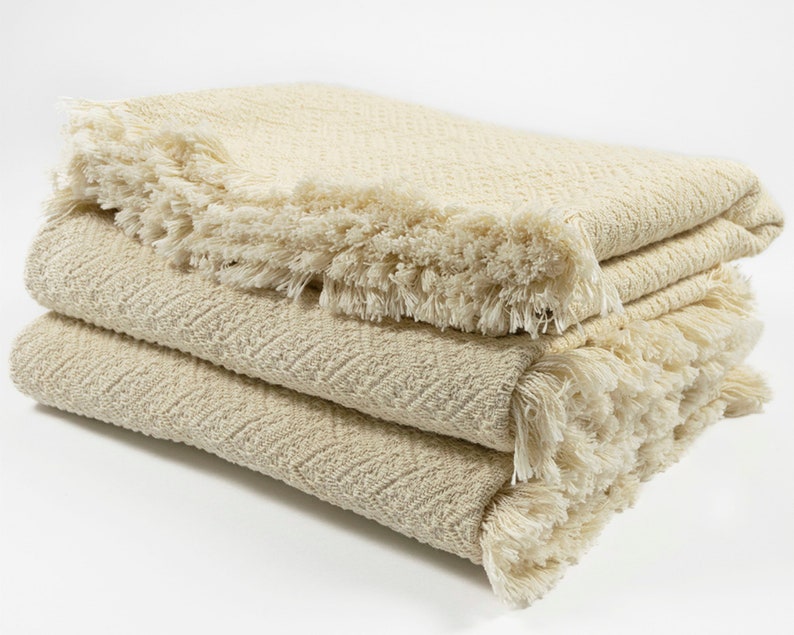 May include: A folded cream-colored throw blanket with a herringbone pattern and fringe trim.