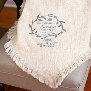 May include: A white throw blanket with a fringed edge and embroidered text that reads "All that we are, All that we HOPE TO BE. we owe to you. Love. Zach & Julie 6.29.2019".