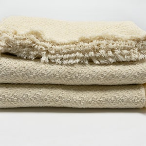 May include: A stack of three cream-colored woven blankets with fringed edges. The blankets are folded and stacked on a white surface.