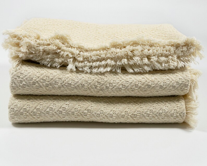 May include: A stack of three cream-colored woven blankets with fringed edges. The blankets are folded and stacked on a white surface.