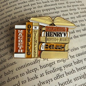 May include: A gold-toned enamel pin shaped like a stack of classic books, including "Macbeth," "Othello," and "Hamlet." An open book sits atop the stack. The pin is set against a backdrop of printed text.