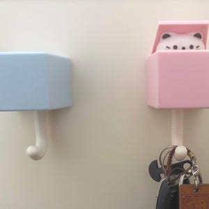 以下が含まれることがあります： 壁掛けフック2つ。1つは水色、もう1つはピンクで猫が覗いています。フックは白で鍵を保持します。フックは装飾的で機能的になるように設計されています。