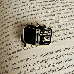 May include: A black enamel pin featuring a cat perched atop a book. The book's cover reads "TO KILL A MOCKINGBIRD" with a gold bird illustration. The pin is set against a backdrop of printed text.