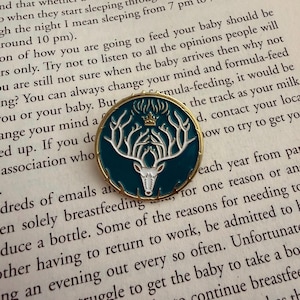 May include: A teal and gold enamel pin with a white stag design, including antlers and a crown. The circular pin is set against a backdrop of printed text. The stag design is detailed and elegant, suitable for a brooch.