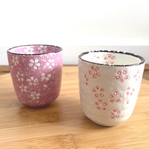 May include: Two ceramic cups with floral patterns. One cup is pink with white flowers, the other is white with pink flowers. Both cups have a black rim and sit on a wooden surface. The cups are approximately 3 inches tall.