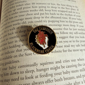 May include: A round black and gold pin with the text "THE THIRTEEN BLACKBEAK COVEN" and a graphic of a heart with red and white details. The pin is on a book page.