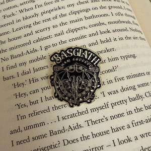 Pode incluir: Um alfinete de esmalte preto e prateado da Basgiath War College, com um design de brasão com um morcego e o texto "BASGIATH WAR COLLEGE". O alfinete está colocado no fundo de um livro aberto.