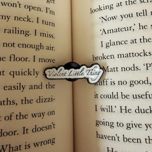 May include: A black and white enamel pin in the shape of a cloud, with the words "Violent Little Thing" in a stylized font. The pin is set against the pages of a book.