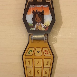 May include: A yellow flip phone toy with a cartoon illustration of a person inside the screen. The phone has a keypad with numbers and symbols. The top of the phone has a decorative design.