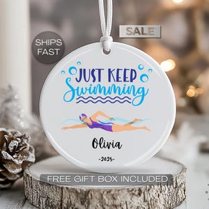 May include: White ceramic ornament with the text "JUST KEEP Swimming" in blue script and a swimmer illustration. The ornament includes the name "Olivia" and the year "-2025-". A white string is attached for hanging.
